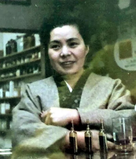 創業者・丸山ミキヱ。薬棚を背にカウンターで腕を組み微笑む
