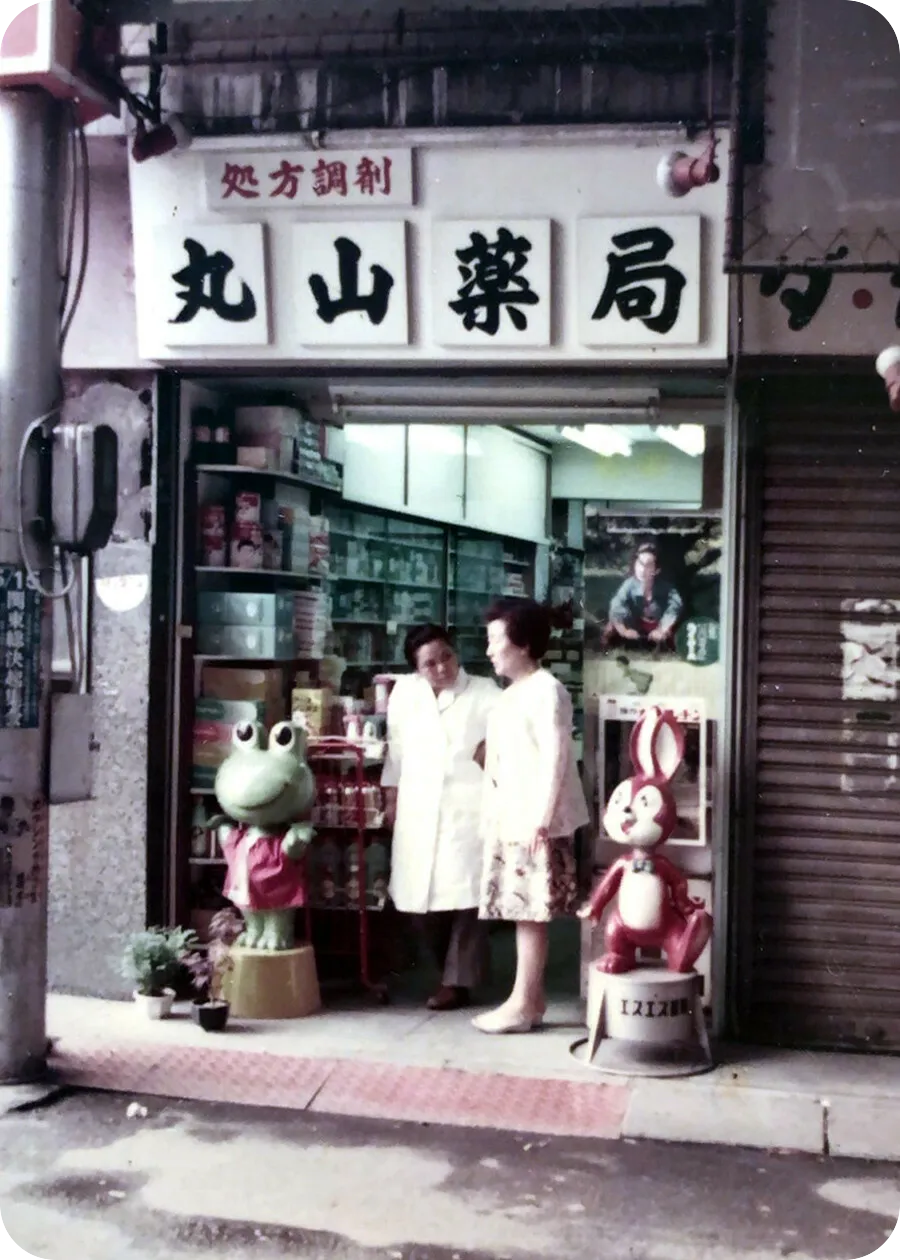 商店街時代の丸山薬局の店頭。白衣の薬剤師と来店客、店先にはカエルとウサギのマスコット人形が並ぶ